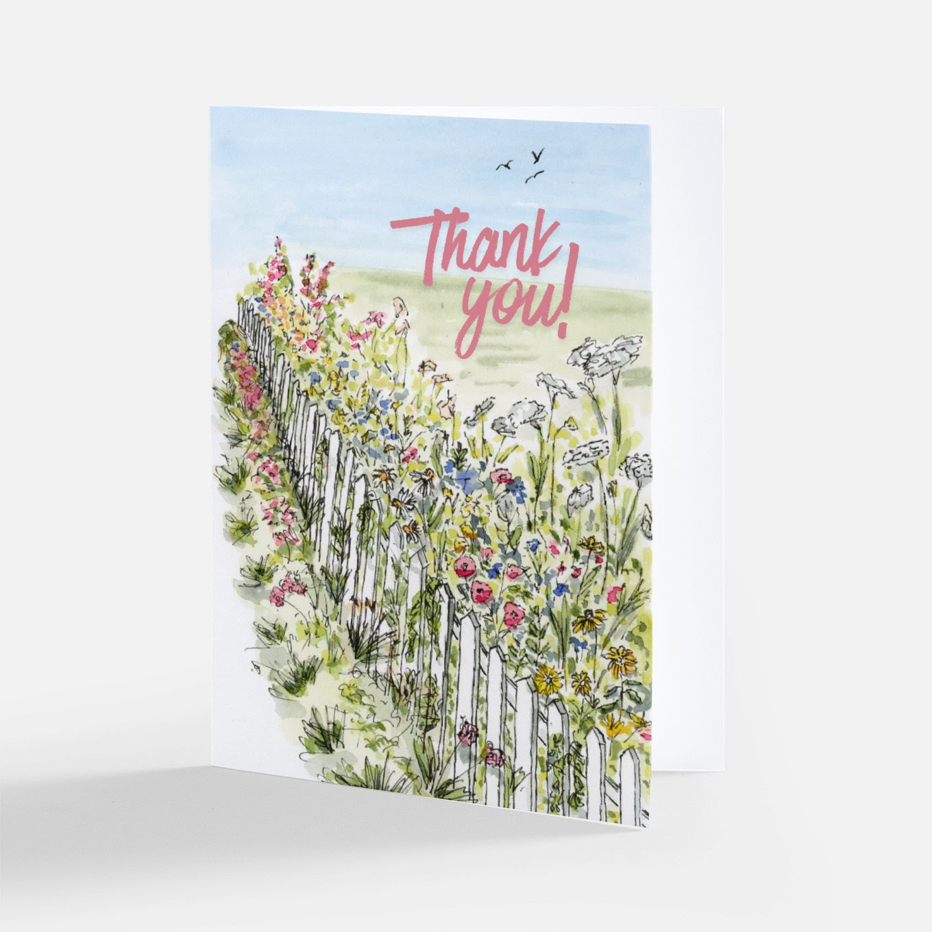 Watercolor thank you card with pink "Thank you!" lettering above a white picket fence lined with pink, blue, and yellow wildflowers and birds in a blue sky